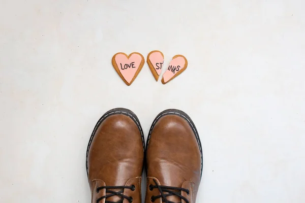 Vista superior de botas de cuero marrón y dos galletas de jengibre corazón roto decoradas con esmalte rosa con el mensaje de amor apesta en el suelo. concepto de angustia . — Foto de Stock