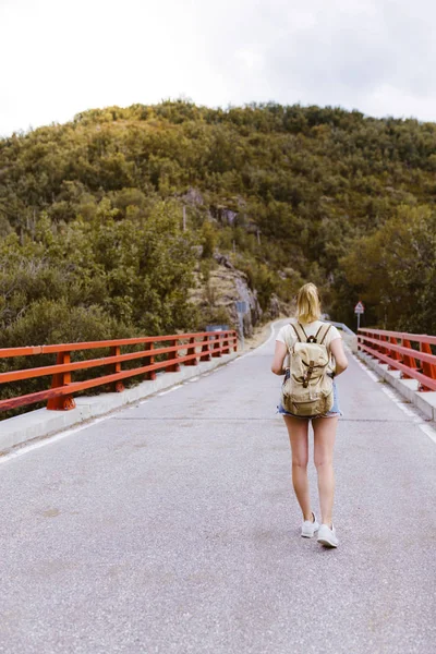 Vista trasera de la joven rubia con mochila caminando por la carretera sobre un puente cerca de la montaña. Concepto de viaje y aventura. Viajero en medio del bosque. Viajar solo — Foto de Stock