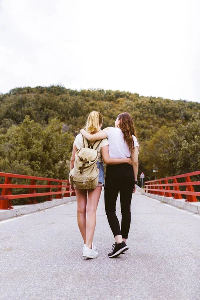 Visszapillantás fiatal barátnők szerelmes hátizsák ölelés az úton, közel a hegyhez. Pár kéz a kézben. Lgtb utazás és kaland koncepció. Utazók az erdő közepén. — Stock Fotó