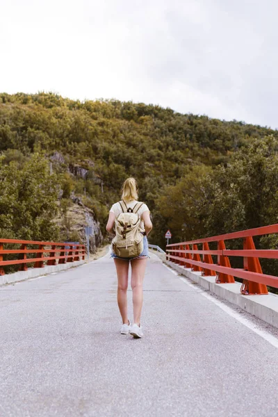 Vista posterior de la joven rubia con mochila caminando por la carretera sobre un puente cerca de la montaña. Concepto de viaje y aventura. Viajero en medio del bosque. Viajar solo — Foto de Stock