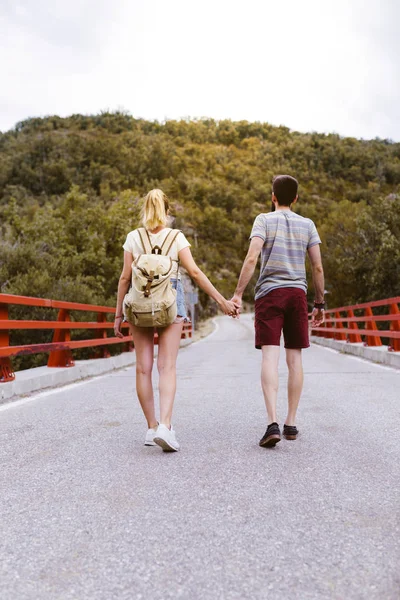 Sırt çantalı genç bir çiftin, dağın yakınındaki bir köprünün üzerinden geçişleri. Aşıklar el ele tutuşuyor. Seyahat ve macera konsepti. Ormanın ortasında gezginler.. — Stok fotoğraf