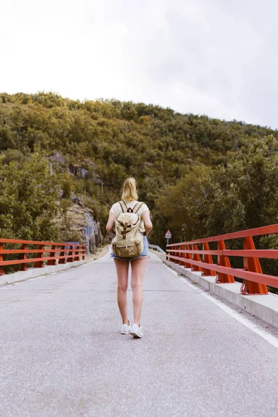 Vue arrière de la jeune femme blonde avec sac à dos marchant sur la route au-dessus d'un pont près de la montagne. Concept voyage et aventure. Voyageur au milieu des bois. Voyager seul Images De Stock Libres De Droits