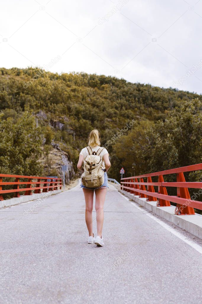 Back view of young blonde woman with backpack walking on the road over a bridge near the mountain. Travel and adventure concept. Traveler in the middle of woodland. Travel alone