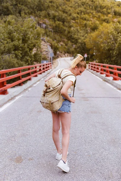 Vista posterior de la joven rubia con mochila caminando por la carretera sobre un puente cerca de la montaña. Concepto de viaje y aventura. Viajero en medio del bosque. Viajar solo — Foto de Stock