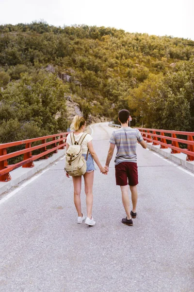 Back view of young couple with backpack walking on the road over a bridge near the mountain. Lovers holding hands. Travel and adventure concept. Travelers in the middle of woodland. — Stock Photo, Image