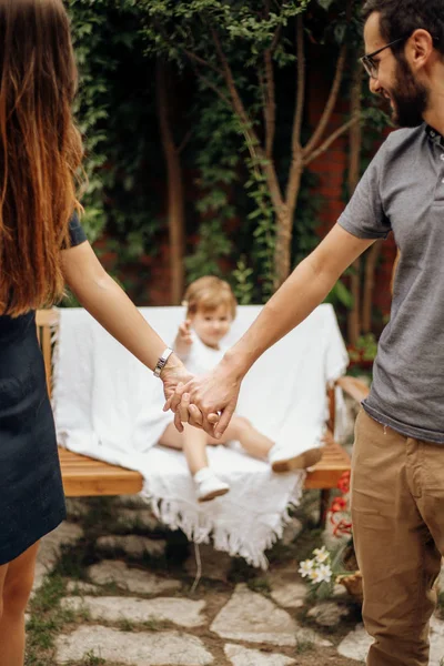Молодые родители держатся за руки со своей маленькой блондинкой на заднем плане в саду. Концепция любви и семьи . Стоковая Картинка