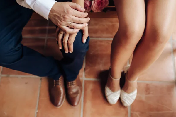 Вид сверху молодоженов с их кольцами. Обувь жениха и невесты и ее маленький свадебный букет из розовых роз сидят и ждут в деревянной скамейке. Концепция дня свадьбы. Селективное фокусирование на кольцах . Стоковое Изображение