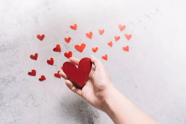 กระดาษแข็งรูปหัวใจสีแดงแวววาวบนมือและหัวใจเล็ก ๆ บนพื้นหลังสีขาว แนวคิดของความรักและวันวาเลนไทน์ กระดาษแข็งรูปหัวใจสีแดงบนมือและหัวใจเล็ก ๆ บนพื้นหลังสีขาว — ภาพถ่ายสต็อก