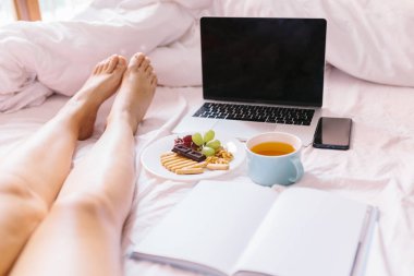 Genç kadın bacakları. Evde yatağında çay, laptopunu kontrol etmek, kitap okumak ve kahvaltı yapmak..