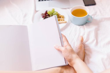 Kitap okuyan genç bir kadın. Evde yatağında çay, laptopunu kontrol etmek, kitap okumak ve kahvaltı yapmak..