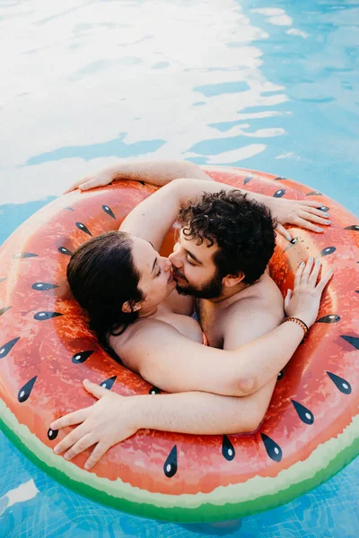 Young couple in love kissing and hugging on watermelon lilo in swimming pool at hotel resort. Summer vacation concept. Love moment.
