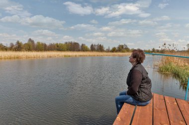 Kafkasyalı orta yaşlı kadın Ros nehrinin kıyısında tahta kaldırımda oturur ve bacaklarına sarılırdı, Ukrayna.