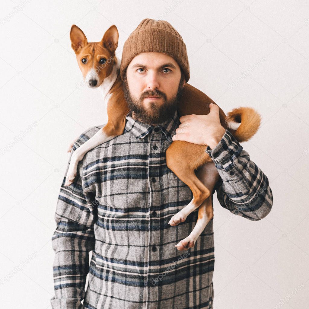 Young white bearded man in a grey plaid shirt and knit hat holding his ginger african basenji dog friend. Friendship between animals and people.