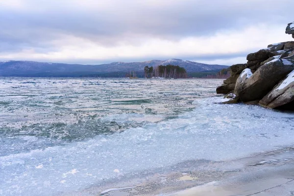 Kış gölünün buzlu kıyısı. Bir orman gölünün kış manzarası. Uzakta alçak dağlar ve ağaçlı bir ada var.