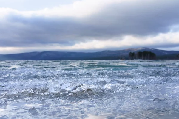 Düşük dağlarla çevrili buzla kaplı göl yüzeyi. Buz kabuğuna zincirlenmiş bir orman gölü kıyısı. Donmuş buz kütleleri ve buz lensleri gölün üzerinde gelişigüzel yatıyor. Uzakta alçak dağlar ve üzerlerinde süzülen kar bulutları görülebilir..