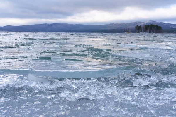 Buzlu orman gölünün kış manzarası. Buz kabuğuna zincirlenmiş bir orman gölünün donmuş kıyısı. Donmuş buz kütleleri ve buz lensleri gölün üzerinde yatıyor. Uzakta alçak dağlar ve kozalaklı ağaçlar görülebilir. Kışın soğuk nefesi.
