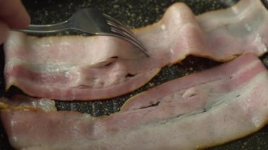 Çatal ızgaranın sıcak yüzeyindeki bir dilim pastırmayı düzeltir. Makronun sıcak tavasında kıtır kıtır domuz pastırması kızartılır, çatal, taş ızgaranın sıcak yüzeyinde bir dilim pastırmayı düzeltir. Yaklaş. Ocakta domuz pastırması şeritleri pişirirken.