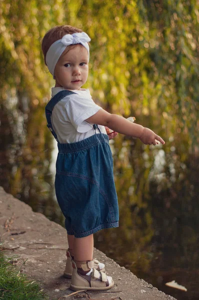Little girl in denim overalls and a white T-shirt feeds geese with bread. — Stock Photo, Image