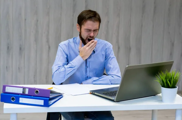 Уставший от скуки молодой бизнесмен, сидящий за столом перед своим компьютером и зевающий — стоковое фото