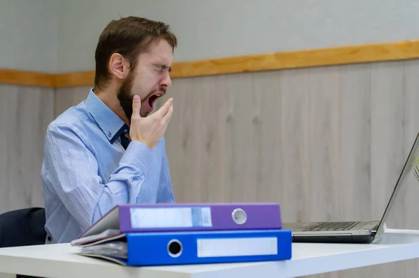 Joven hombre de negocios aburrido y cansado sentado en su escritorio frente a su computadora bostezando — Foto de Stock