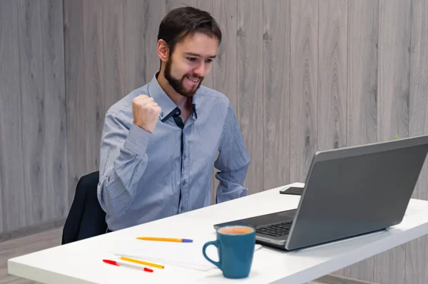 Vaya, es una gran victoria. Joven hombre de negocios sentado en la oficina, mirando la pantalla del ordenador portátil y sonriendo, apriete los puños saborear algunas buenas noticias, ganar, lograr la meta —  Fotos de Stock