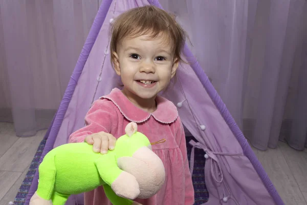 Little girl 2 years old smiling and show colorful toys near wigwam in childrens room. — Stock Photo, Image