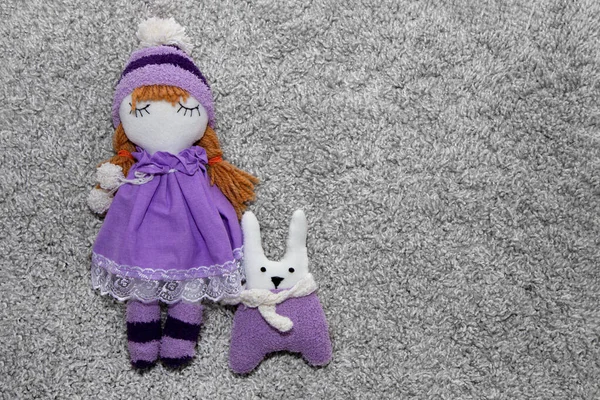 Handmade doll in purple hat and dress with toy bunny on gray background