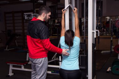 Kişisel antrenörü olan bir kadın, kablolu ağırlık makinesinde spor salonunda barfiks çekiyor.