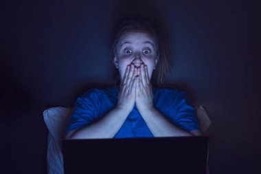 Kız geceleri yatakta oturuyor ve korkmuş bir şekilde dizüstü bilgisayara bakıyor. mavi ışık