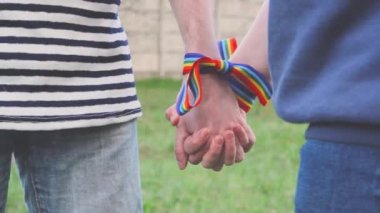 Bileklerinde gökkuşağı LGBT kurdeleleri olan eller. Eşcinsel çift nazikçe el ele tutuşup birbirlerini okşuyor.