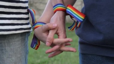 Bileklerinde gökkuşağı LGBT kurdeleleri olan eller. Eşcinsel çift nazikçe el ele tutuşup birbirlerini okşuyor.