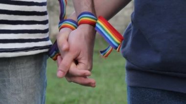 Bileklerinde gökkuşağı LGBT kurdeleleri olan eller. Eşcinsel çift nazikçe el ele tutuşup birbirlerini okşuyor.