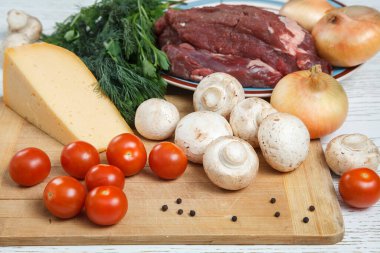 Yemek yapmak için gerekli malzemeler. Tahta üzerinde çiğ et, soğan, kiraz domatesleri, yeşiller, domatesler, peynir, otlar, baharatlar.