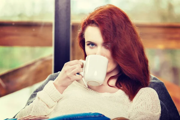 Atractiva mujer pelirroja bebiendo una taza de café sentada en la mecedora. Chica joven con bebida energizante caliente —  Fotos de Stock