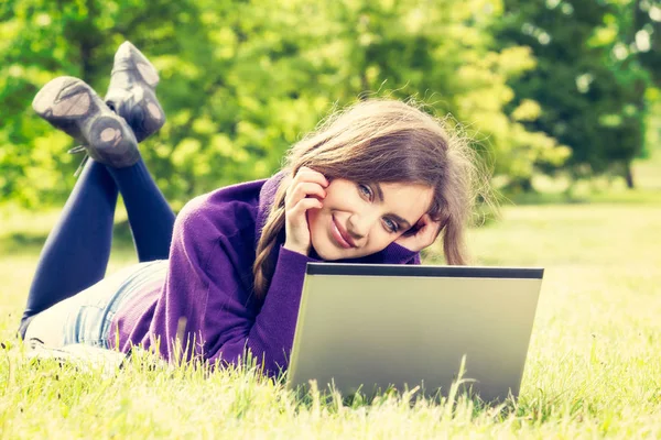 Mujer joven usando el ordenador portátil en el parque acostado en la hierba verde — Foto de Stock