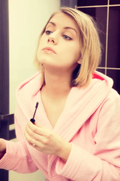 Blonde woman wearing in dressing-gown applying mascara in mirror at home — Stock Photo, Image