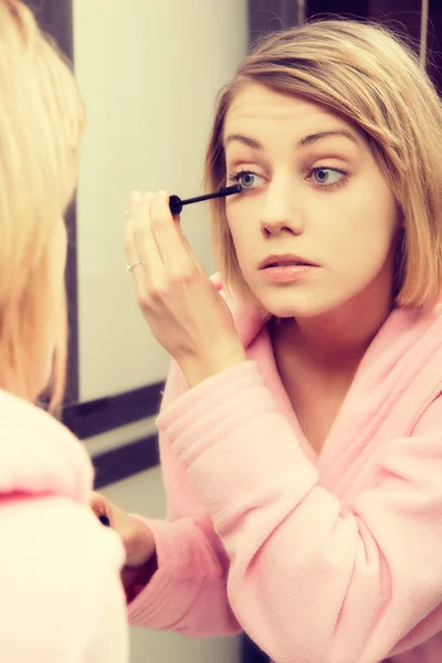 Blonde woman wearing in dressing-gown applying mascara in mirror at home — Stock Photo, Image
