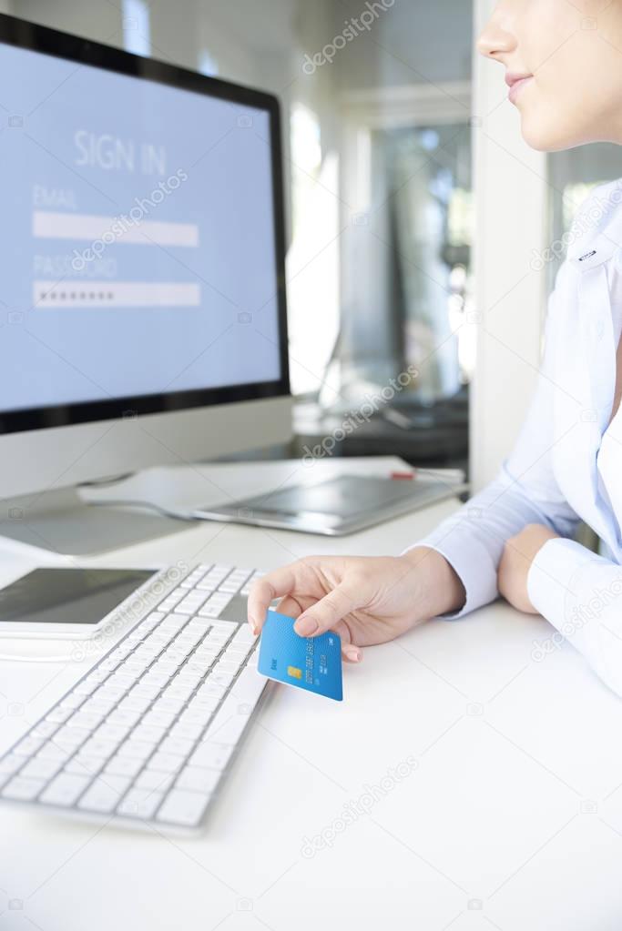 Close-up shot of a young businesswoman sitting in the office in front of computer and holding credit card while signing  in.
