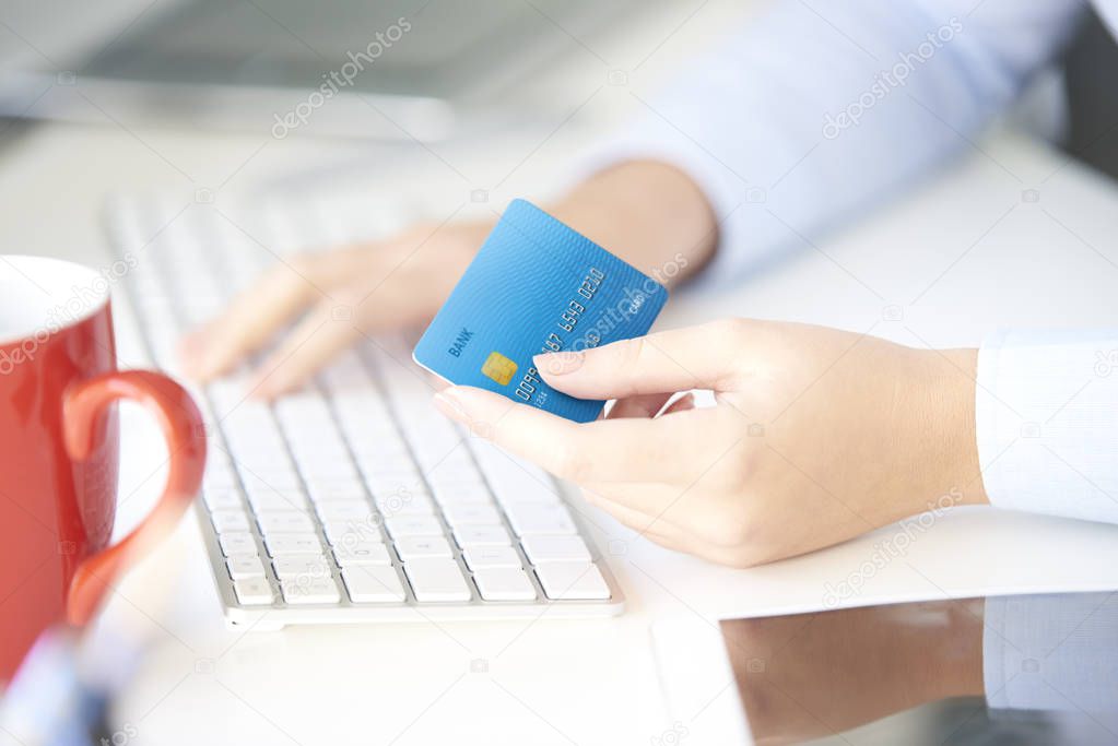 Close-up shot of a young businesswoman sitting in the office in front of computer and holding credit card while typing data.