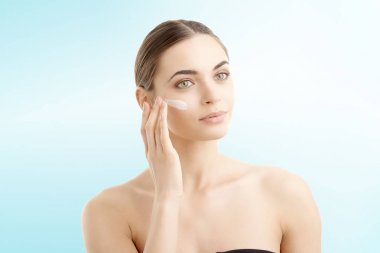 Studio shot of beautiful young woman applying moisturizer onto her face against at isolated light blue background with copy space.  clipart