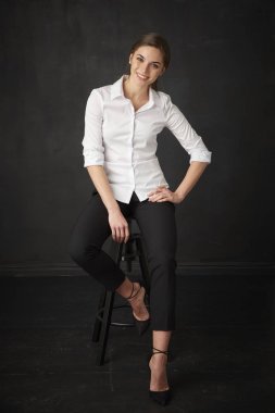 Kameraya bakarak ve koyu arka plan otururken gülümseyen güzel genç kadının tam uzunlukta studio vurdu. 