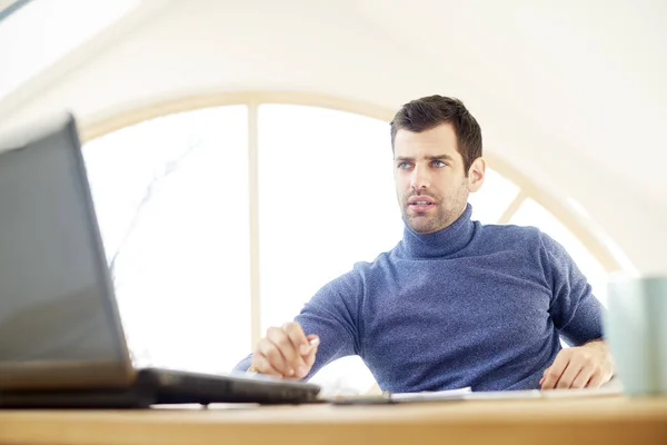 在家里使用笔记本电脑时 穿着高领毛衣和看 Thoughtul 的休闲年轻人的肖像 家庭办公室 — 图库照片
