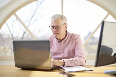 Gömlek giymiş ve laptop evden çalışırken masa başında oturan emekli adam. 