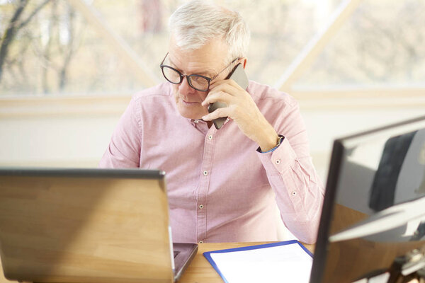 Высокоуровневый снимок пожилого бизнесмена, сидящего перед ноутбуком и разговаривающего с кем-то на мобильном телефоне во время работы
. 