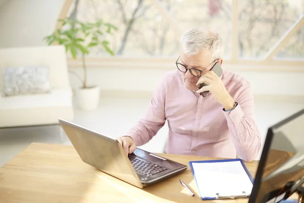 High angle shot of elderly businessman sitting in front of laptop and and talking with somebody on his mobile phone while working hard.