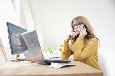 Cep telefonu kullanarak ve istemci ile konuşurken, ofis büro dizüstü bilgisayar önünde oturmak ve onun iş yönetme orta yaşlı Danışmanı iş kadını. 