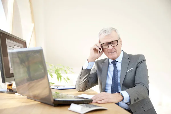 Portrait of a senior sales man sitting at office desk in front of laptop and talking with somebody in his mobile phone.