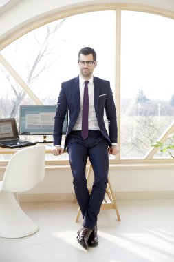 Takım elbise ve ofis masa başında otururken kameraya bakarak yakışıklı genç işadamı doz tam uzunlukta. 