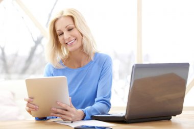 Dizüstü bilgisayar önünde masada oturan ve çevrimiçi çalışıyorsanız dijital tablet kullanarak rahat iş kadını gülümseyen. Ev ofis. 