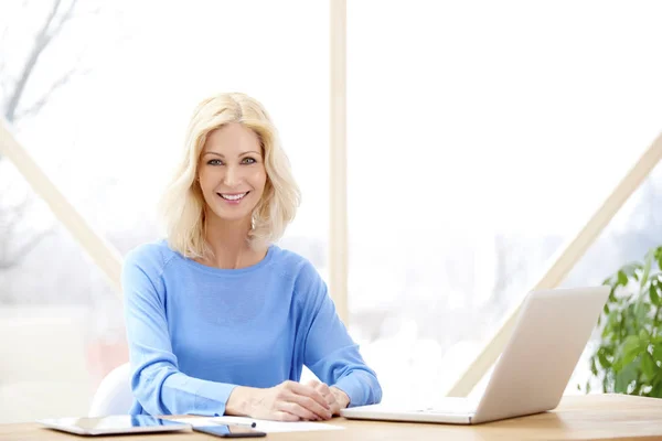 照片中的美丽中年妇女看着相机和微笑 而坐在办公桌前的笔记本电脑 家庭办公室 — 图库照片
